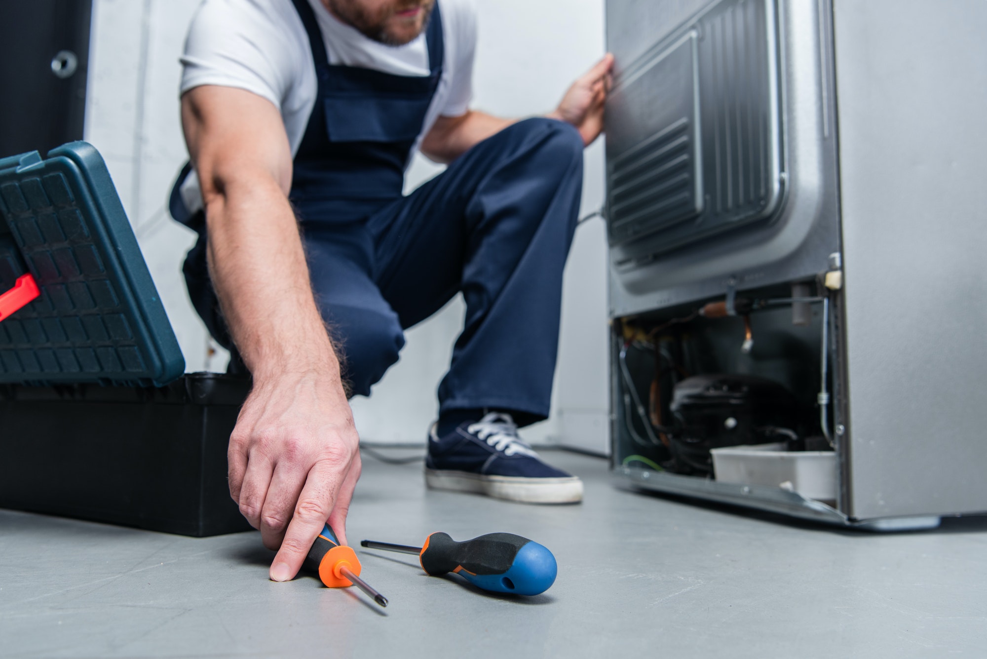 cropped-shot-of-repairman-in-working-overall-taking-screwdriver-from-floor-near-broken-refrigerator